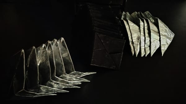 Twelve matte black parallelograms folded such that they have three segments rest in a pile flanked on either side by a pile of six reflective parallelograms similarly folded.